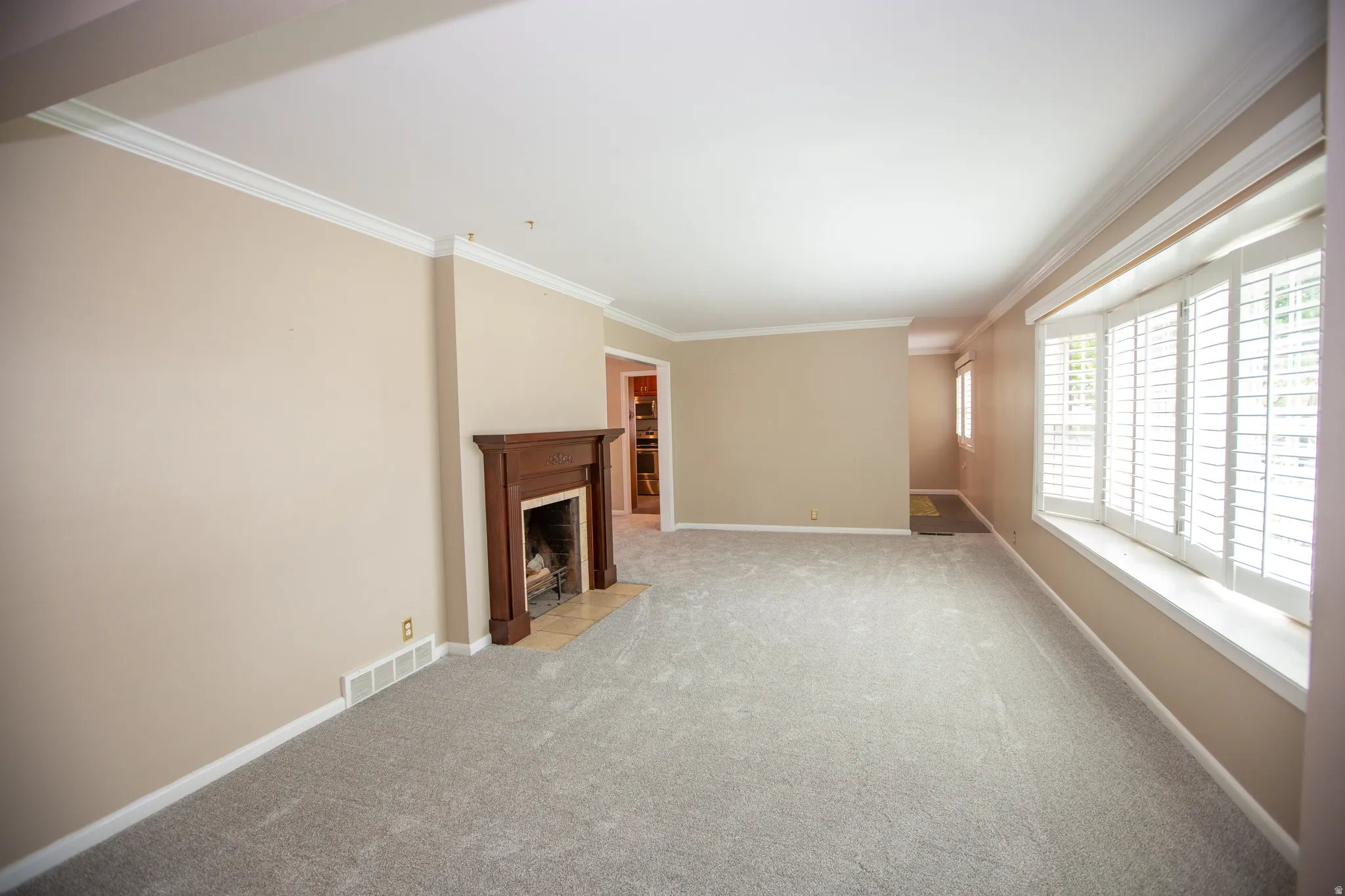 Spacious living area featuring a wood mantel fireplace with tile hearth, plush carpeting, crown molding, and large windows with plantation shutters