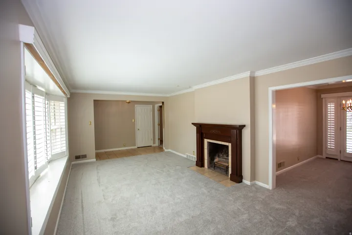 Spacious room featuring light gray carpeting, a wood-surround fireplace with a hearth, a bay window with plantation shutters, crown molding, and a doorway opening to another room