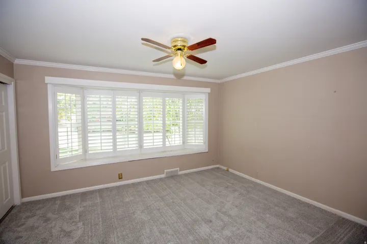 Room featuring a prominent bay window with white plantation shutters, crown molding, a ceiling fan with light fixture, neutral wall paint, and gray carpet flooring