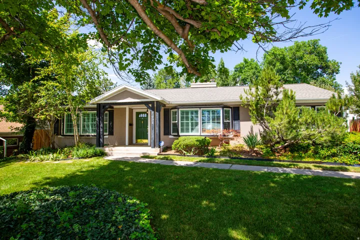 Ranch-style residence featuring a covered entry, dark shutters, a green front door, a gray shingle roof, and a mature landscaped front yard with a manicured lawn