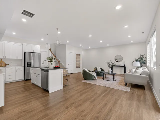 Open-concept living space featuring wood-finish flooring throughout, a modern kitchen with white cabinetry and stainless steel appliances, a center island with a farm sink, recessed lighting, and a staircase with natural wood treads