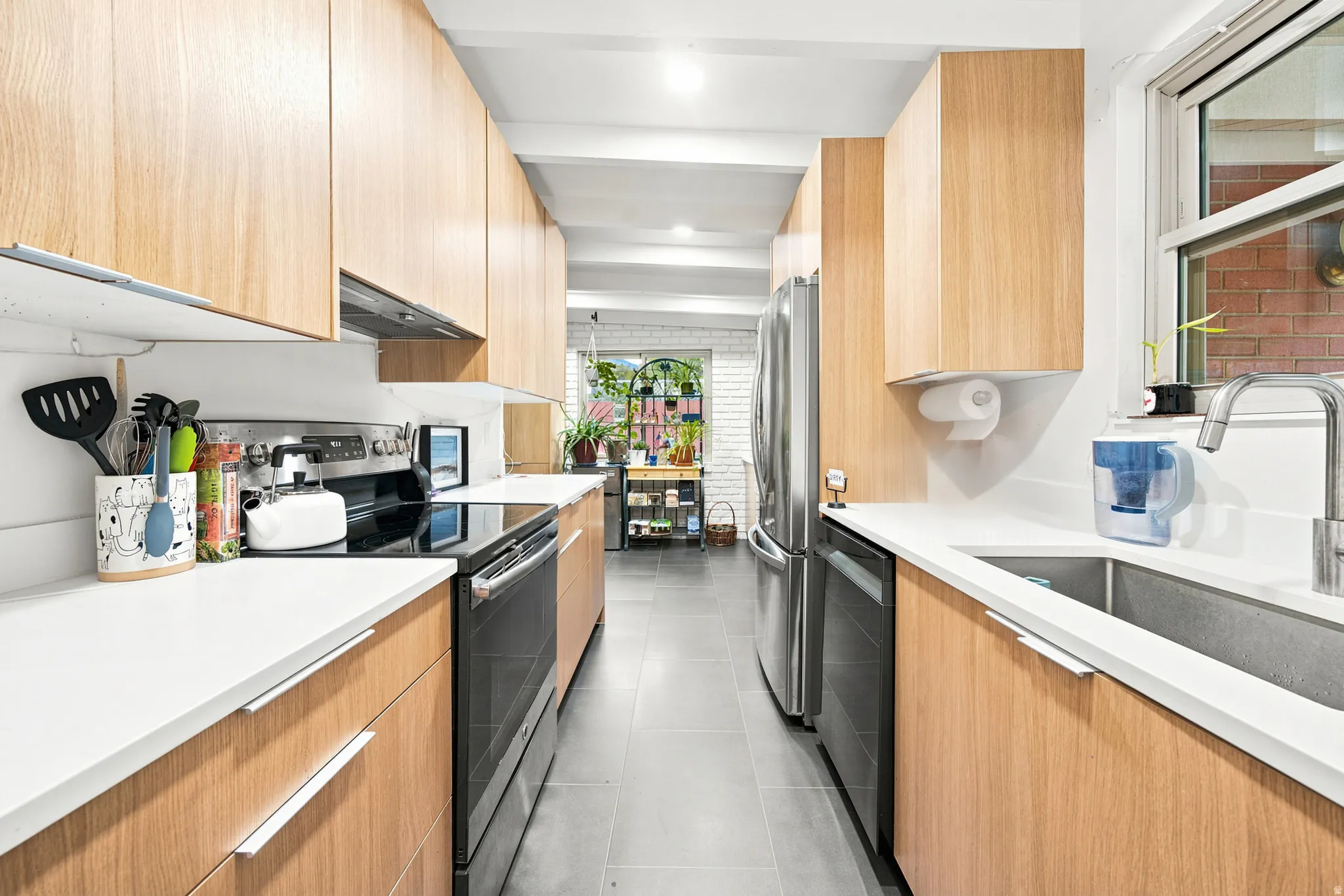 Galley kitchen featuring light wood-finish cabinetry, white solid-surface countertops, stainless steel appliances, and a stainless steel sink with a gooseneck faucet