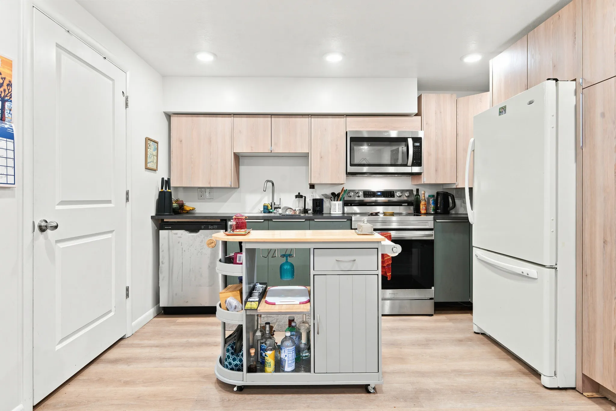 Kitchen featuring light wood-finish cabinetry, stainless steel appliances, dark countertops, and wood-finish flooring