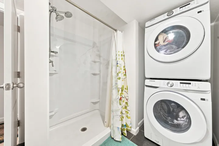 Compact bathroom featuring a shower with built-in shelving, a dual showerhead, and a stacked washer and dryer unit