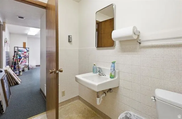 Restroom featuring a wall-mounted sink with chrome faucet, a rectangular mirror, white ceramic wall tile, and a single-flush toilet