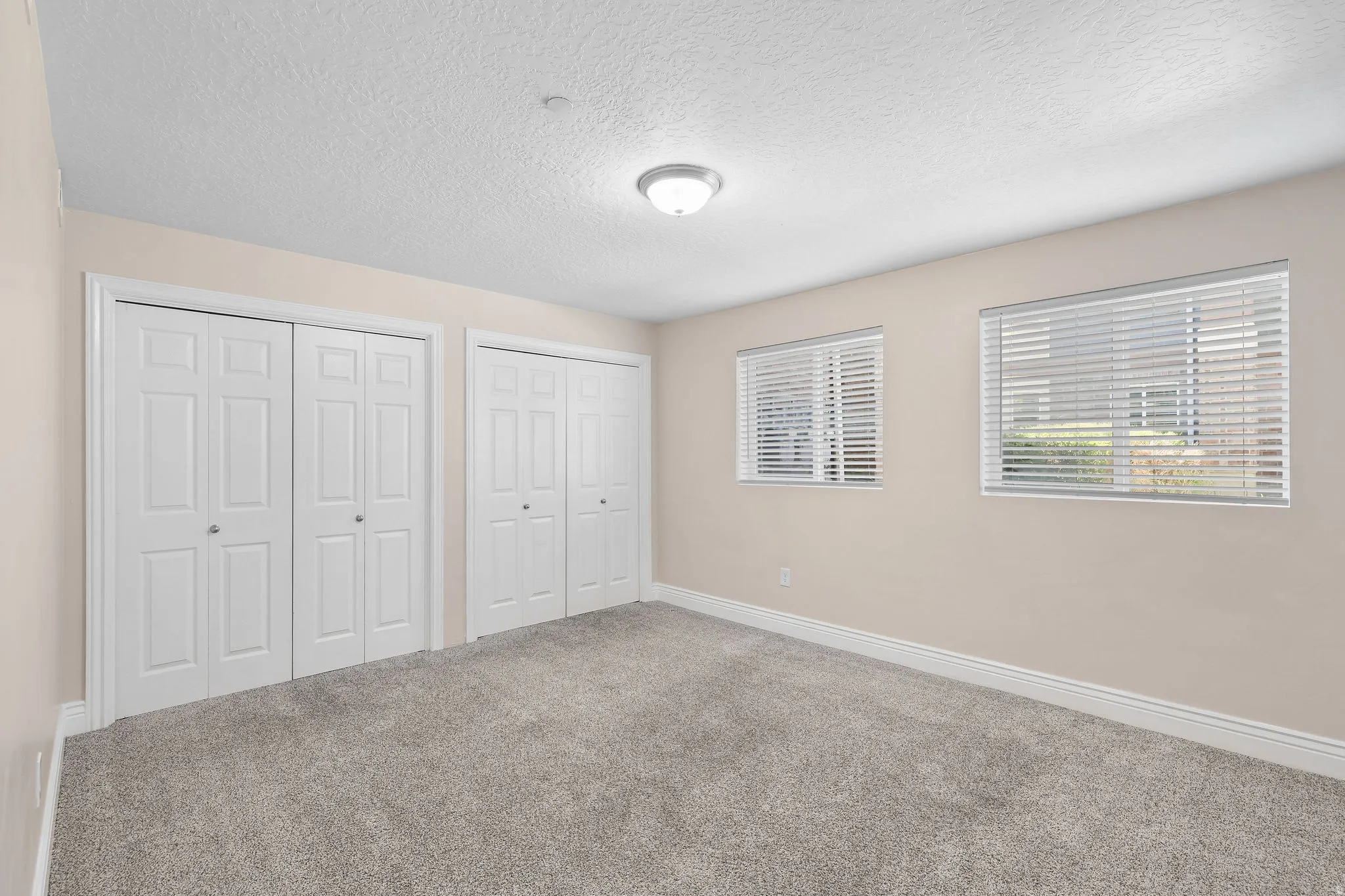 Room featuring two sets of bi-fold closet doors, two windows with horizontal blinds, neutral-toned walls, and grey carpet flooring