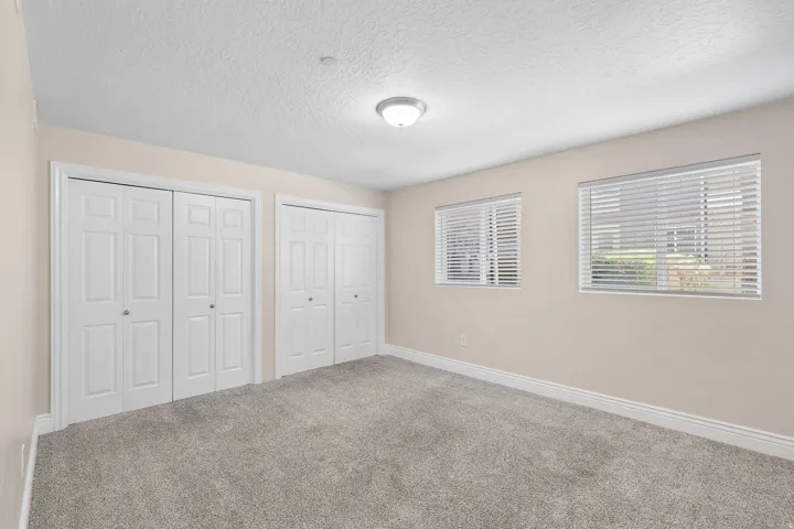 Room featuring two sets of bi-fold closet doors, two windows with horizontal blinds, neutral-toned walls, and grey carpet flooring