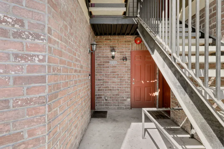 Brick exterior featuring a red entry door, sconce lighting, concrete walkway, and a metal staircase with railing