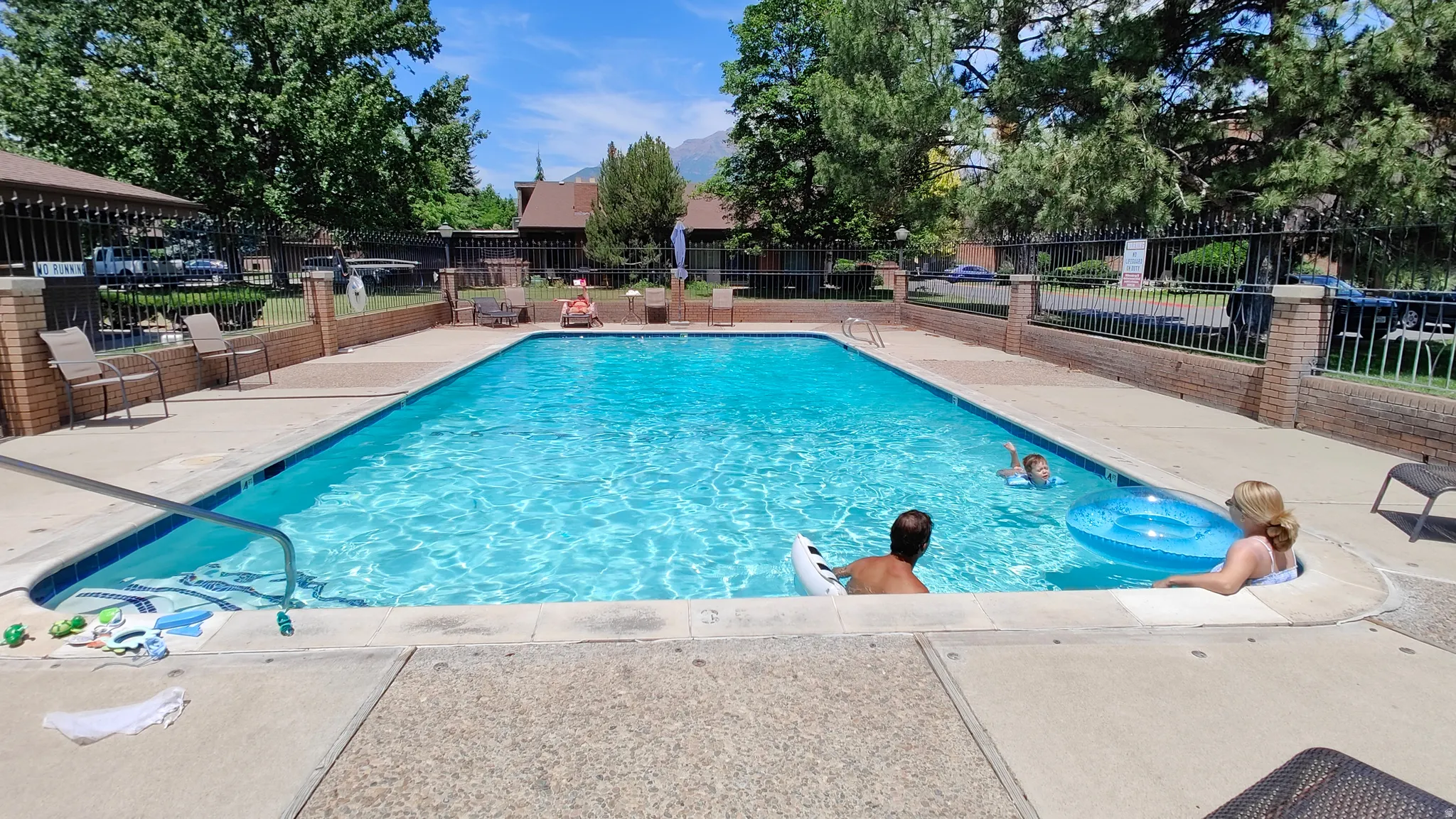 Rectangular outdoor swimming pool featuring concrete decking, perimeter fencing with brick pillars, and mature trees providing shade