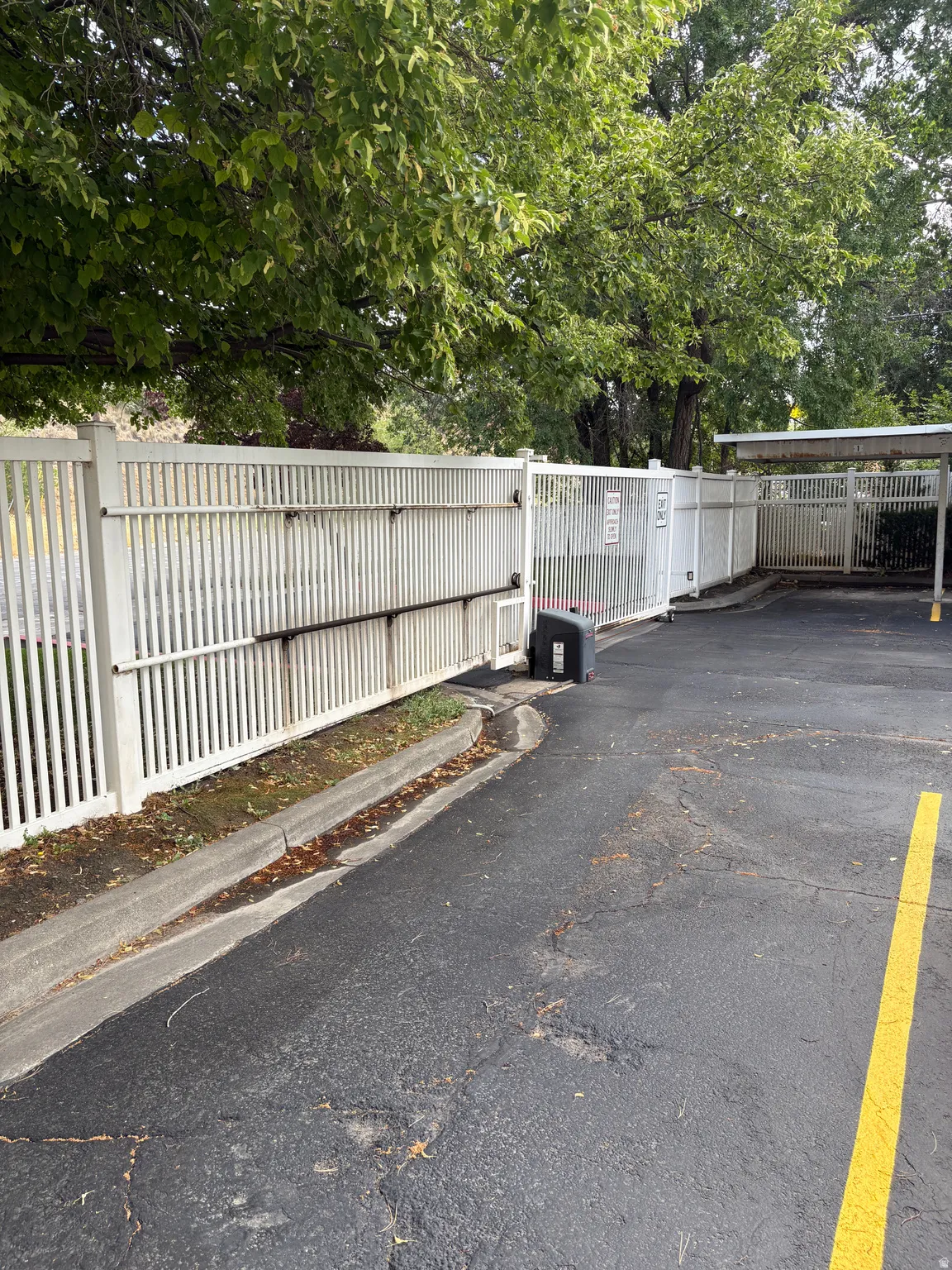 Paved asphalt parking area featuring a white picket fence, mature trees, and a covered parking structure