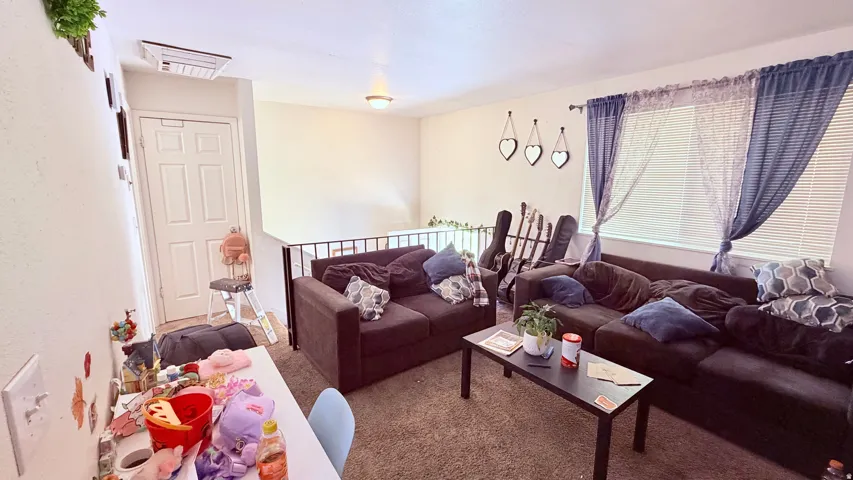 Carpeted living area featuring a privacy window with blinds, a wall-mounted AC unit, and a metal railing overlooking the lower level