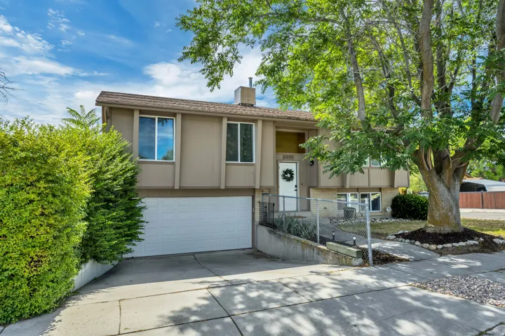 Split-level exterior featuring a two-car garage, mature front yard trees, established shrubbery, and a concrete driveway with a sidewalk
