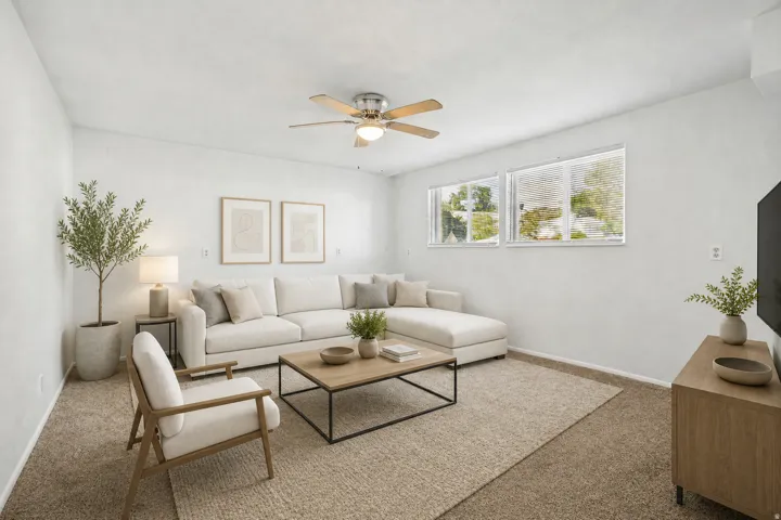 Bright living space featuring neutral carpeting, a ceiling fan with light, and multiple windows providing natural illumination