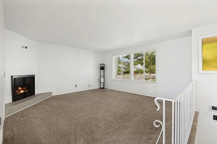 Living area featuring a corner fireplace with a raised hearth, broad windows, neutral carpet flooring, and a decorative white railing
