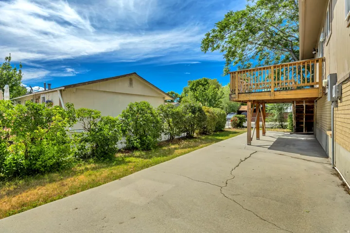Rear exterior featuring a raised wood deck, mature trees, concrete patio, and established shrubbery