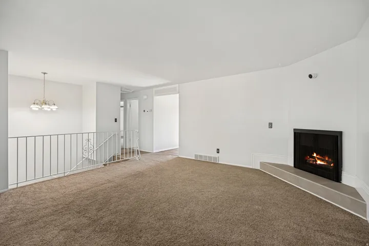 Living area featuring a corner fireplace with a built-in hearth, textured carpeting, white painted walls, a metal stair railing, and an overhead chandelier
