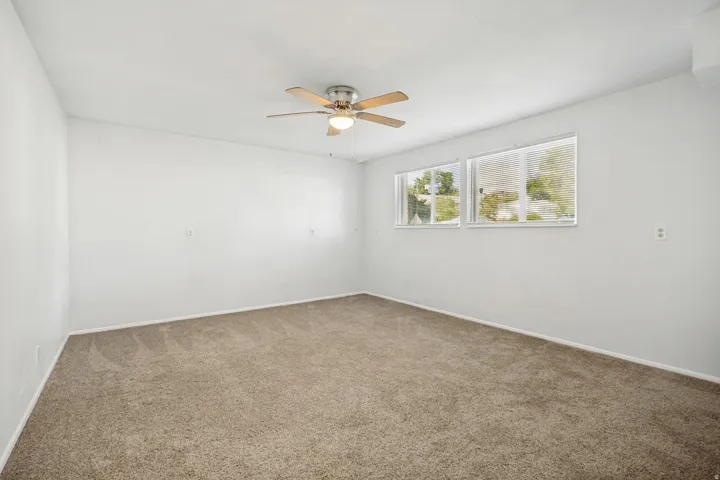 Spacious room featuring neutral carpeting, white walls, two horizontal windows, and a ceiling fan with light fixture