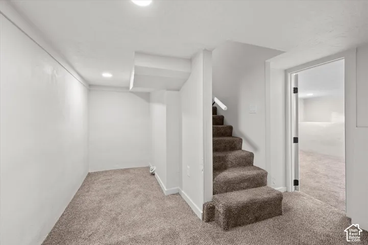 Carpeted interior space featuring a staircase with a white handrail, recessed lighting, white walls, and a doorway opening to an additional room