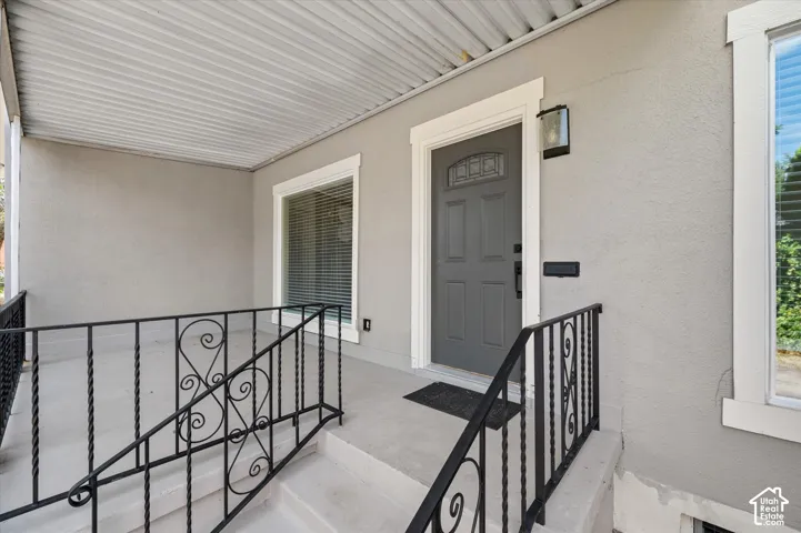 Covered entry porch featuring a paneled entry door with decorative glass, an exterior light fixture, a window with white trim, and a black wrought iron handrail