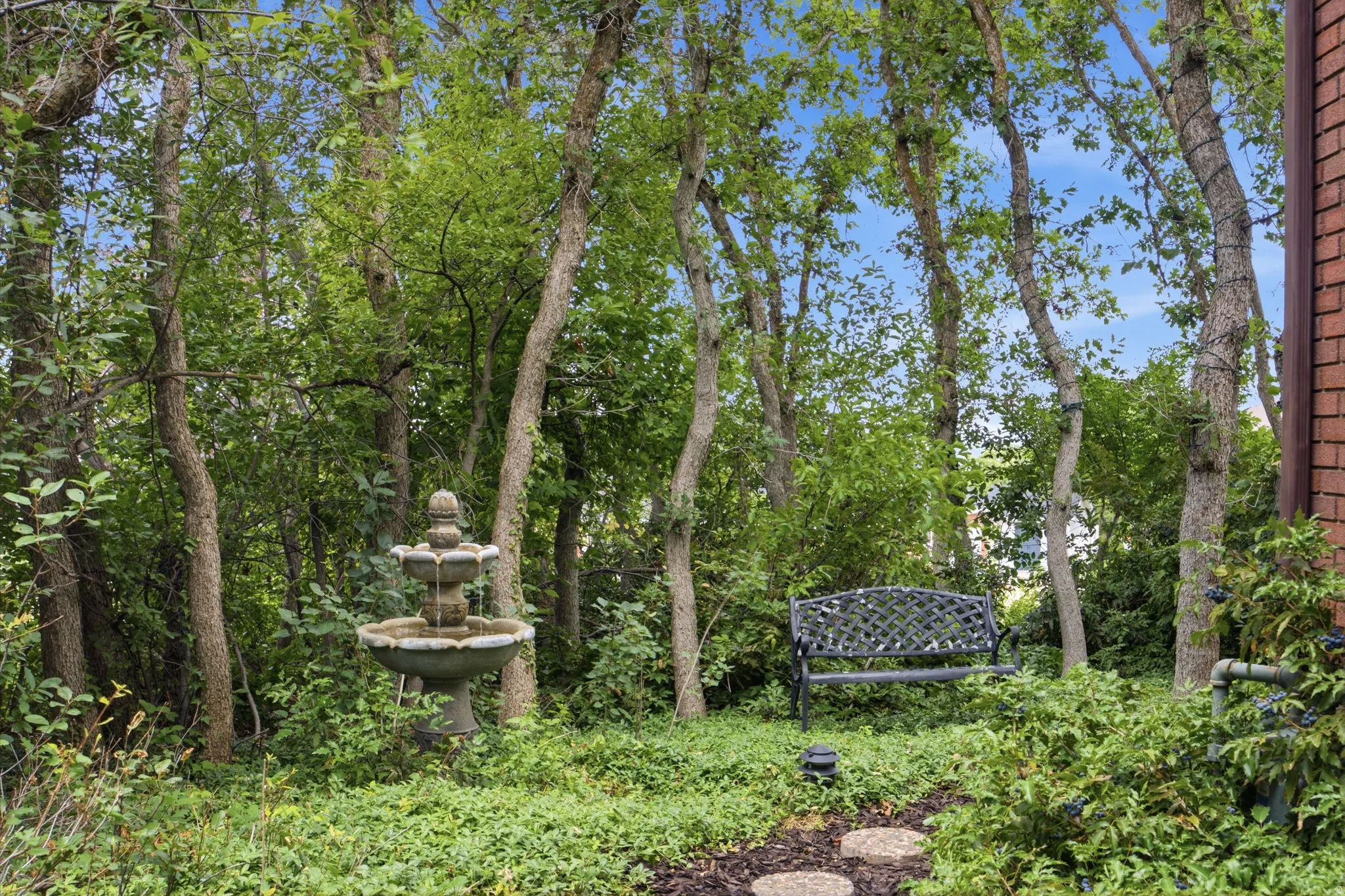 Landscaped garden area featuring mature trees, a multi-tiered stone fountain, and a decorative metal bench