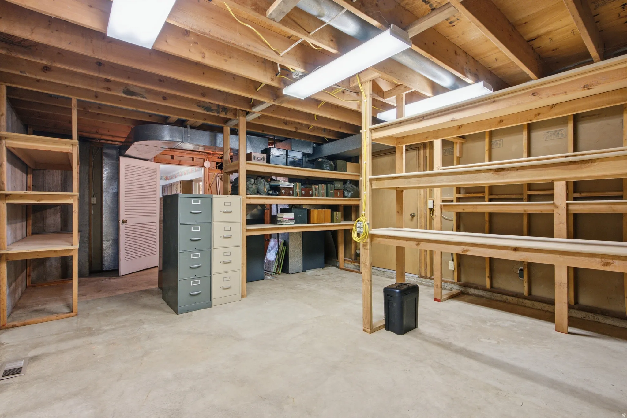 Unfinished basement featuring extensive wood shelving, a workshop bench, concrete flooring, exposed ceiling joists, and fluorescent overhead lighting