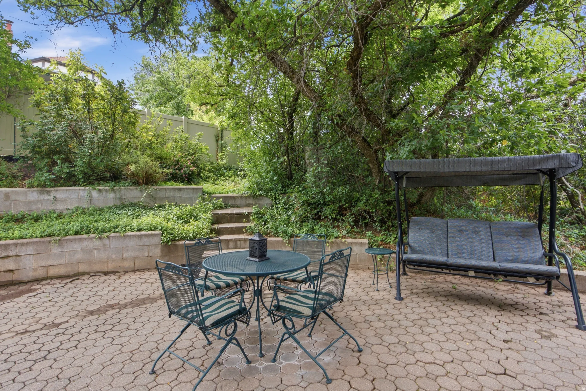 Paved patio featuring a geometric paver design, surrounded by mature trees and layered garden beds with concrete retaining walls
