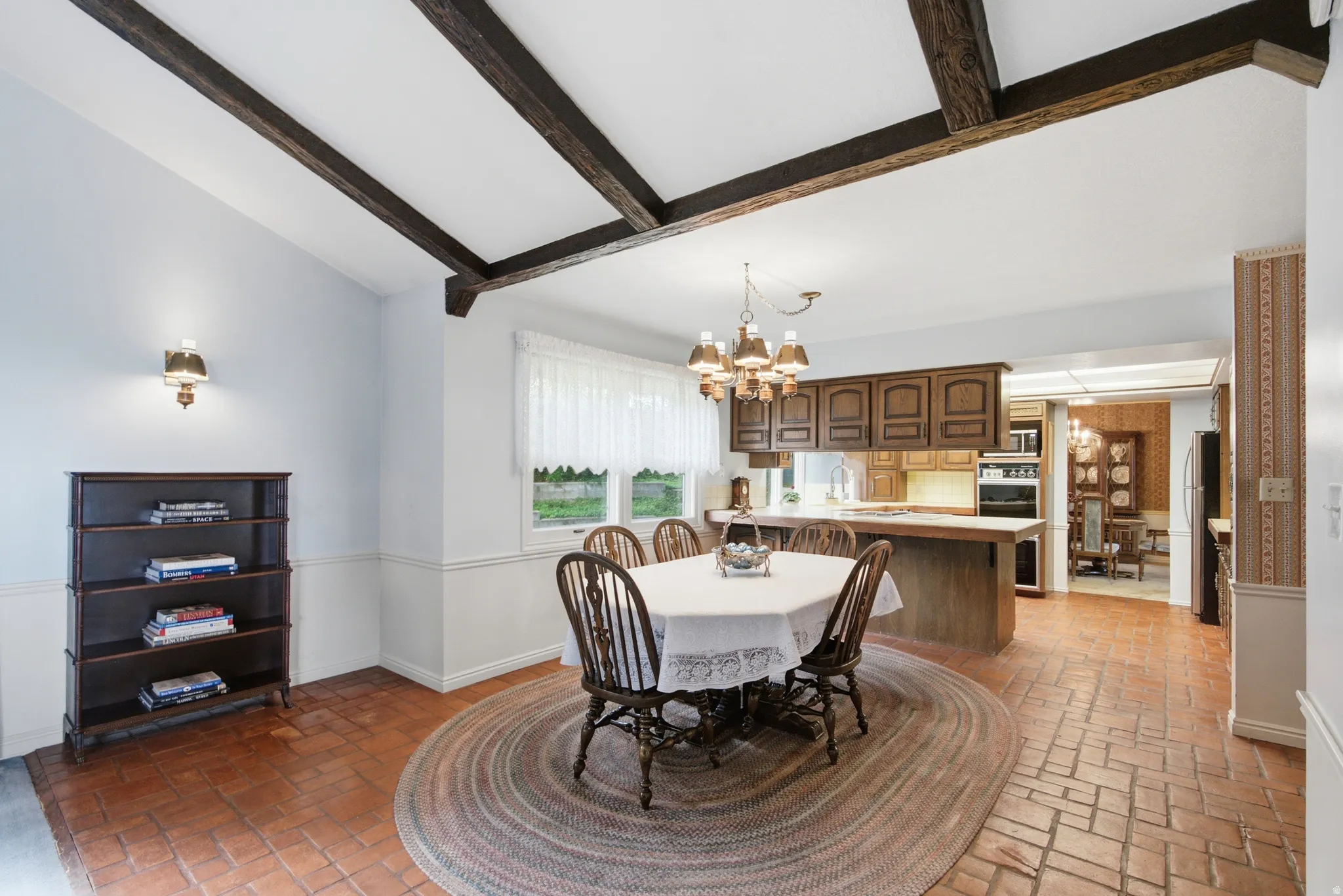 Vaulted ceiling with exposed wood beams, brick-finish flooring, wood cabinetry, built-in wall oven, and a chandelier fixture