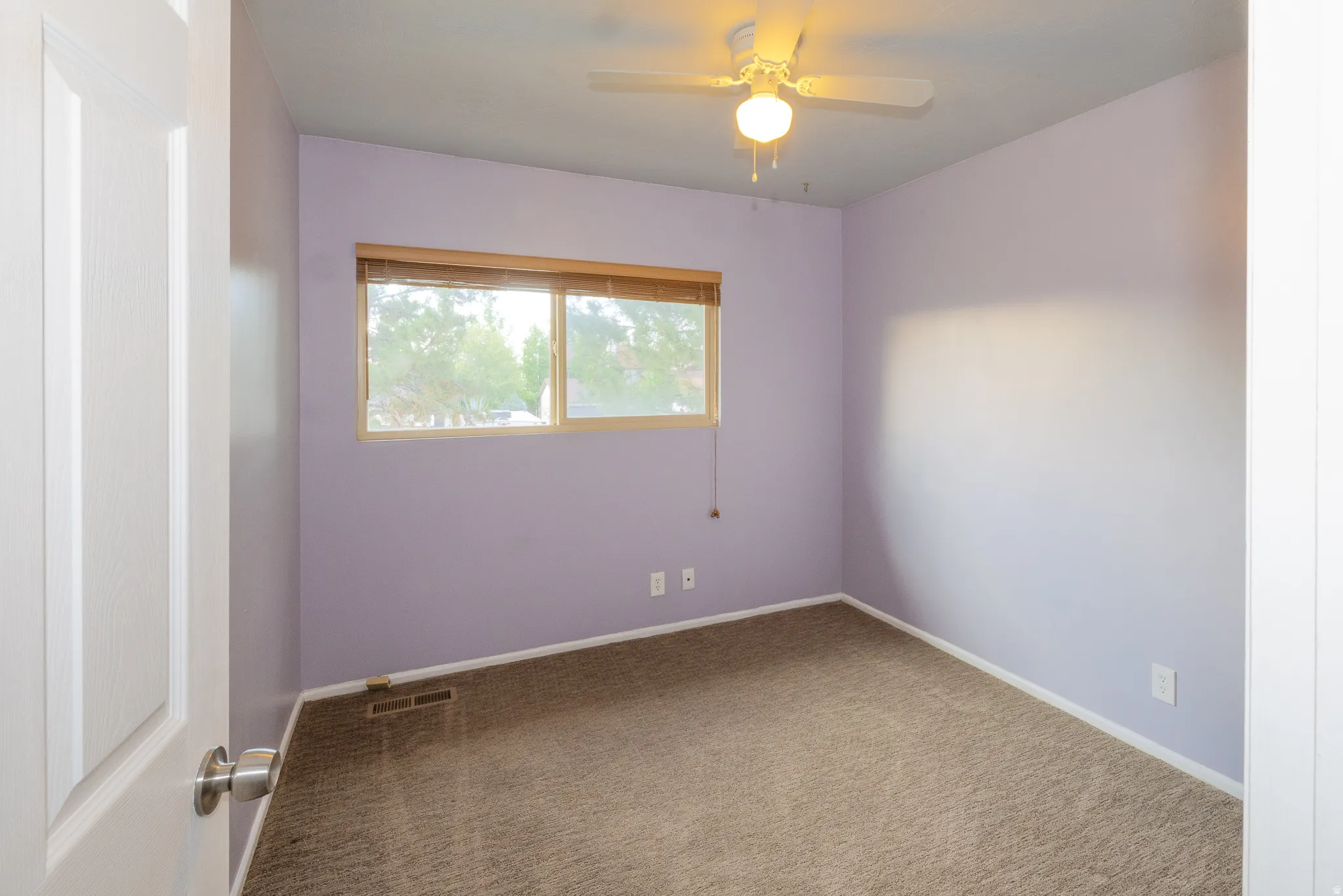 Room with light purple walls, brown carpet, and a window with wood-finish blinds