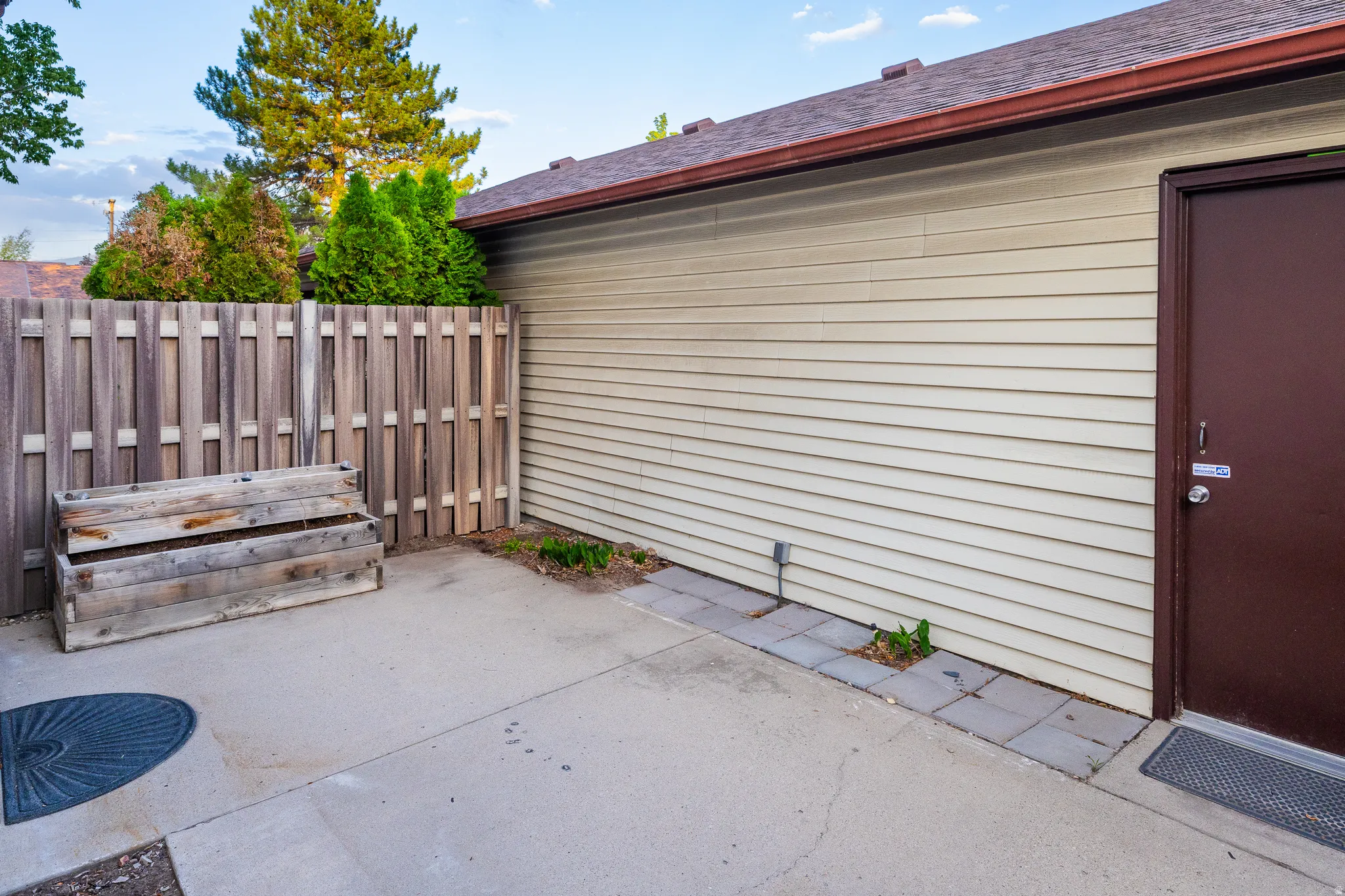 Concrete patio area featuring a privacy fence, wood planter box, light-toned siding, brown exterior door, and a matching brown roofline