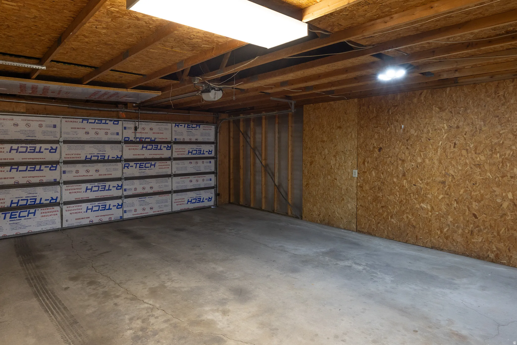 Spacious garage featuring a concrete floor, exposed wood ceiling joists, and a garage door with R-TECH insulation