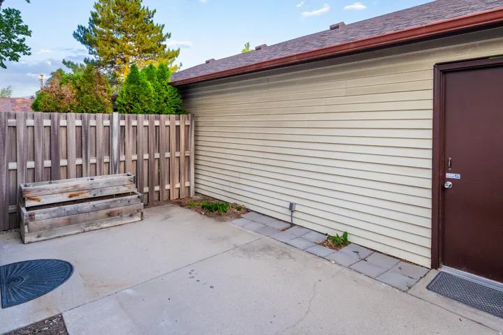 Concrete patio area featuring a privacy fence, wood planter box, light-toned siding, brown exterior door, and a matching brown roofline