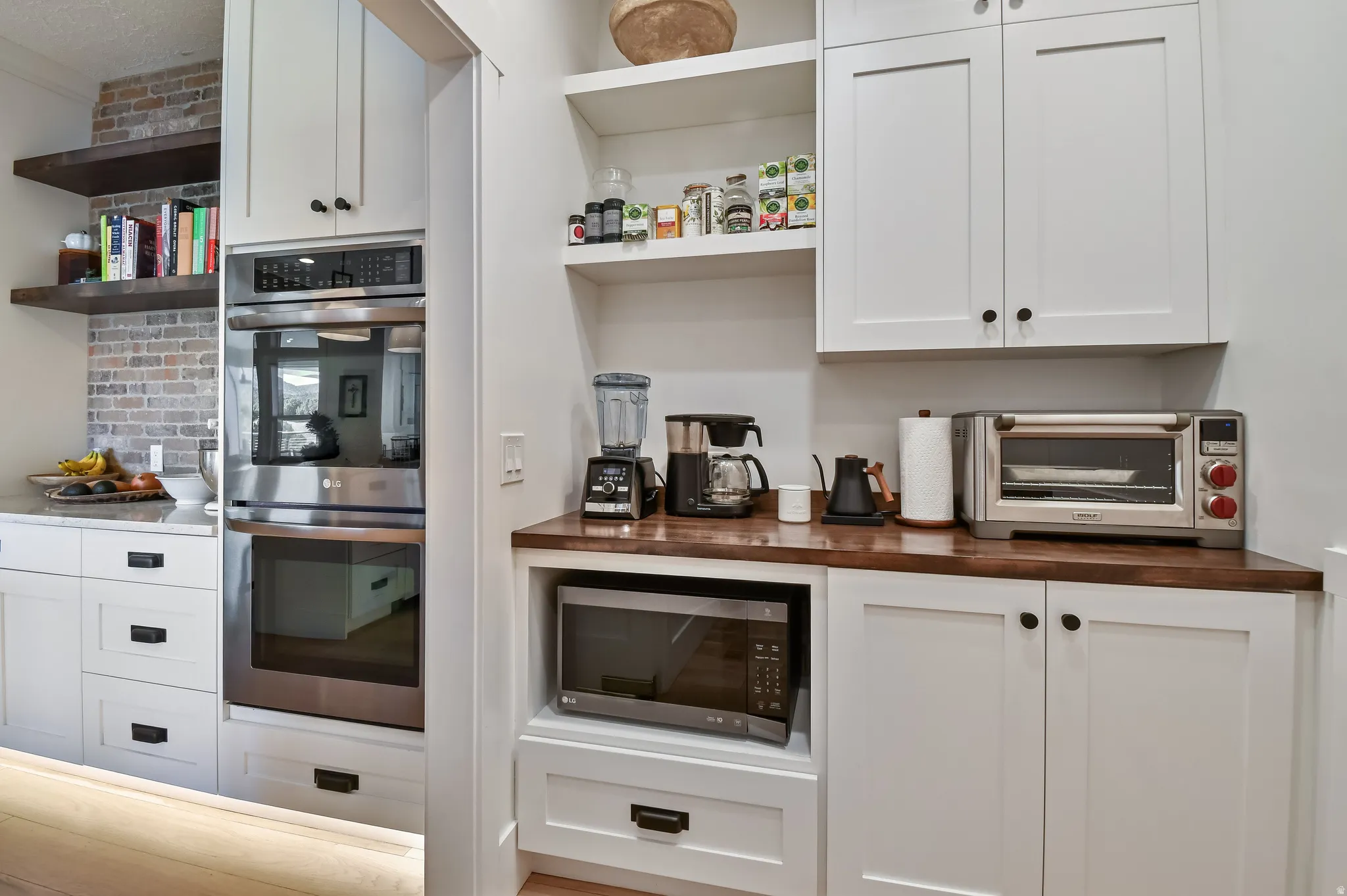Kitchen features white shaker cabinetry with dark hardware, built-in stainless steel oven and microwave, and open shelving with brick-look tile backsplash