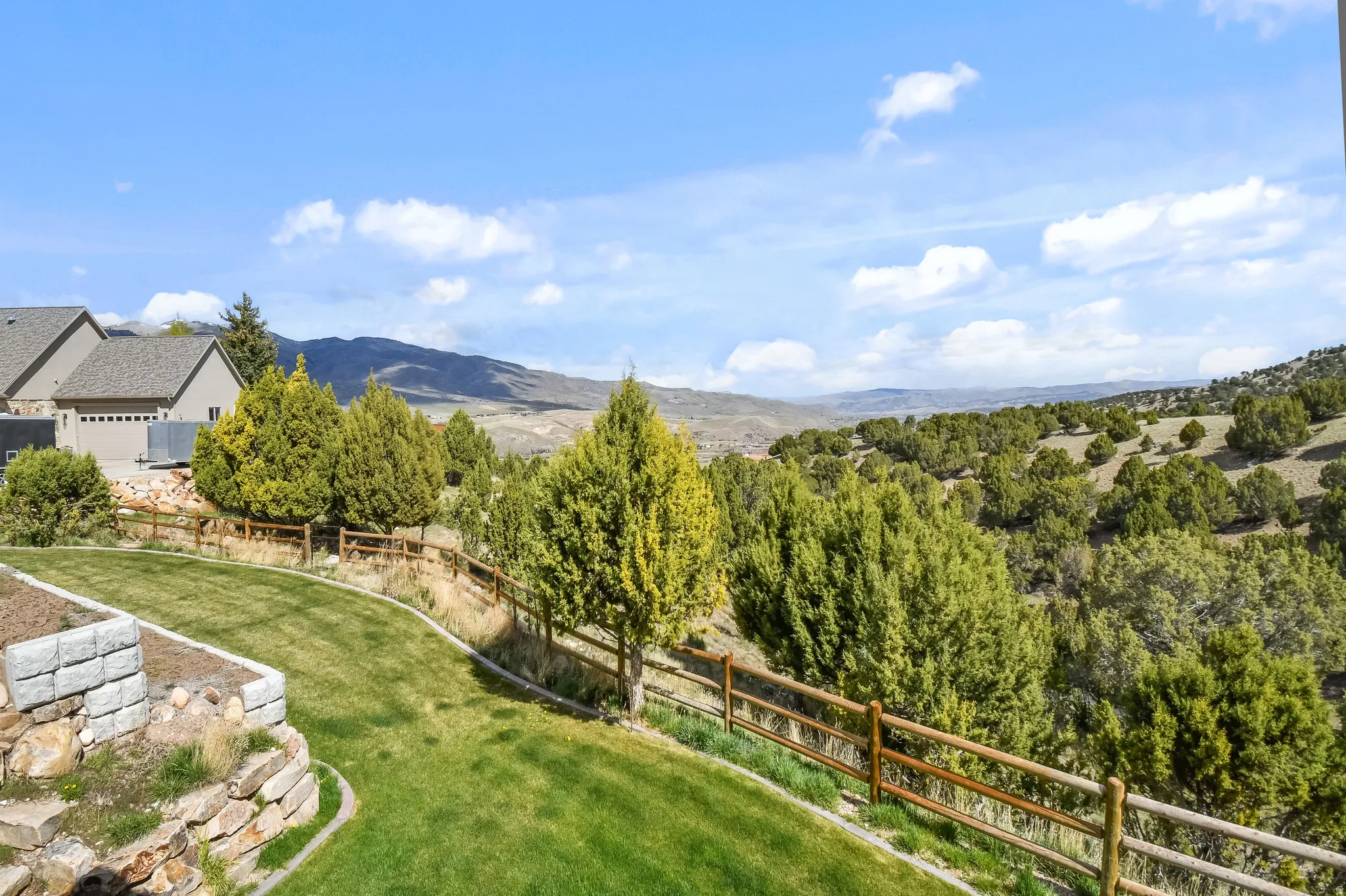 Expansive backyard featuring a manicured lawn, tiered landscaping with stone retaining walls, and a rustic split-rail fence bordering natural terrain
