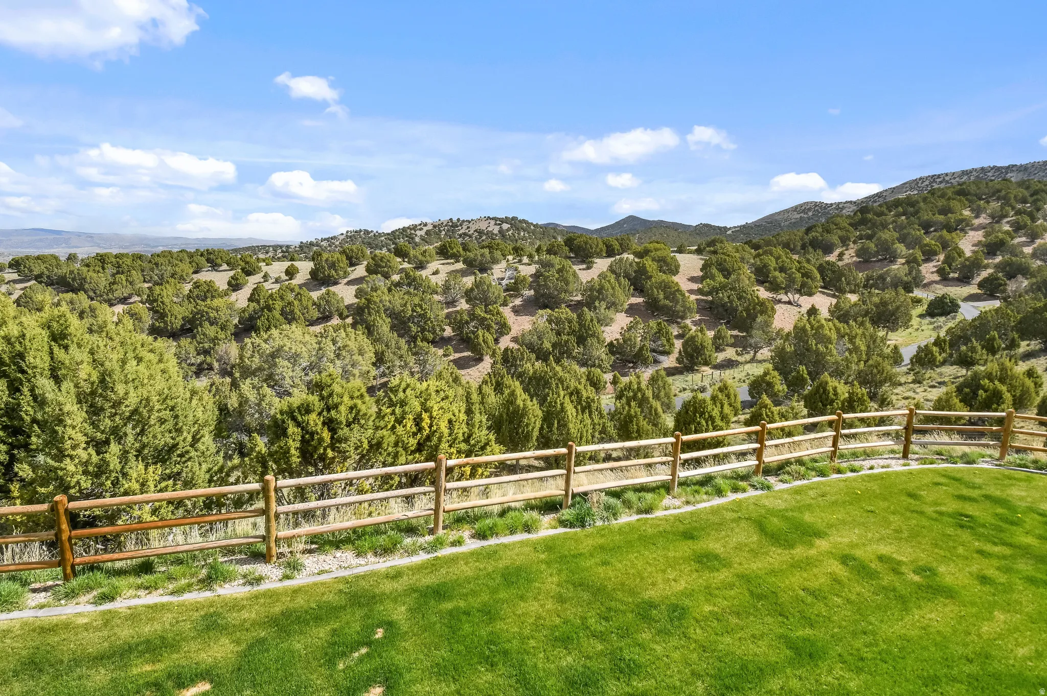 Expansive outdoor area featuring a manicured green lawn, a rustic wooden rail fence, and a natural landscape with rolling hills and abundant trees under a clear sky