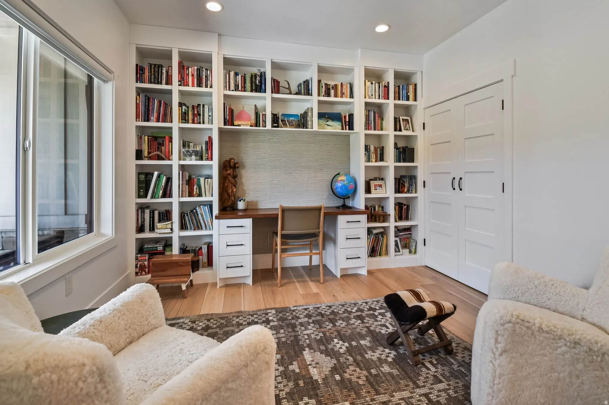 Built-in white shelving system with integrated desk, hardwood flooring, large window, recessed lighting, and white paneled doors