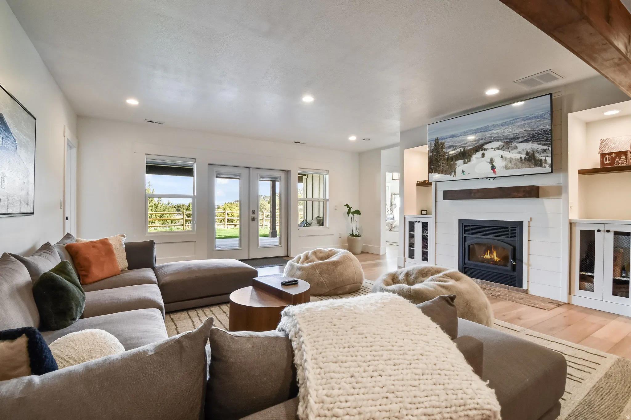Basement family room with spacious living area featuring exposed wood beam, recessed lighting, wood-finish flooring, a shiplap fireplace with a dark metal insert, and rear-yard access via glass-paned double doors