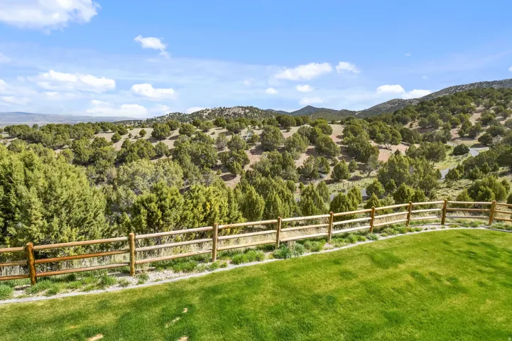 Expansive outdoor area featuring a manicured green lawn, a rustic wooden rail fence, and a natural landscape with rolling hills and abundant trees under a clear sky