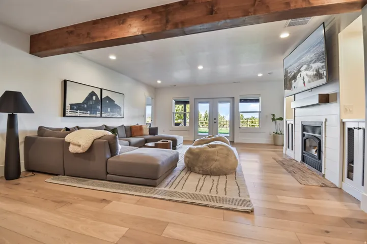 Basement family room with spacious living area featuring exposed wood beam, recessed lighting, wood-finish flooring, a shiplap fireplace with a dark metal insert, and rear-yard access via glass-paned double doors