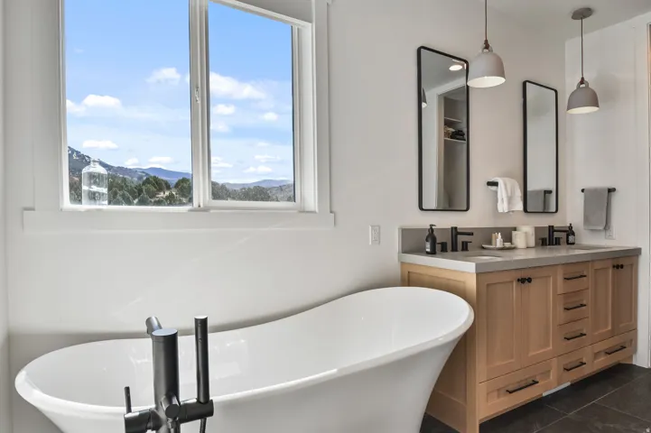 Primary bathroom with freestanding soaking tub with a floor-mounted fixture, dual vanity with light wood cabinetry, stone countertop, undermount sinks, and matte black fixtures