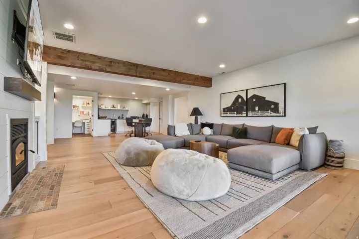 Basement family room with spacious living area featuring exposed wood beam, recessed lighting, wood-finish flooring, a shiplap fireplace with a dark metal insert, and rear-yard access via glass-paned double doors