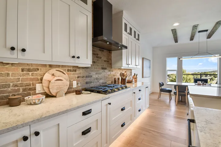 Modern kitchen featuring white shaker cabinetry with matte black hardware, brick backsplash, under-cabinet lighting, natural wood-finish flooring, and a window with a distant landscape
