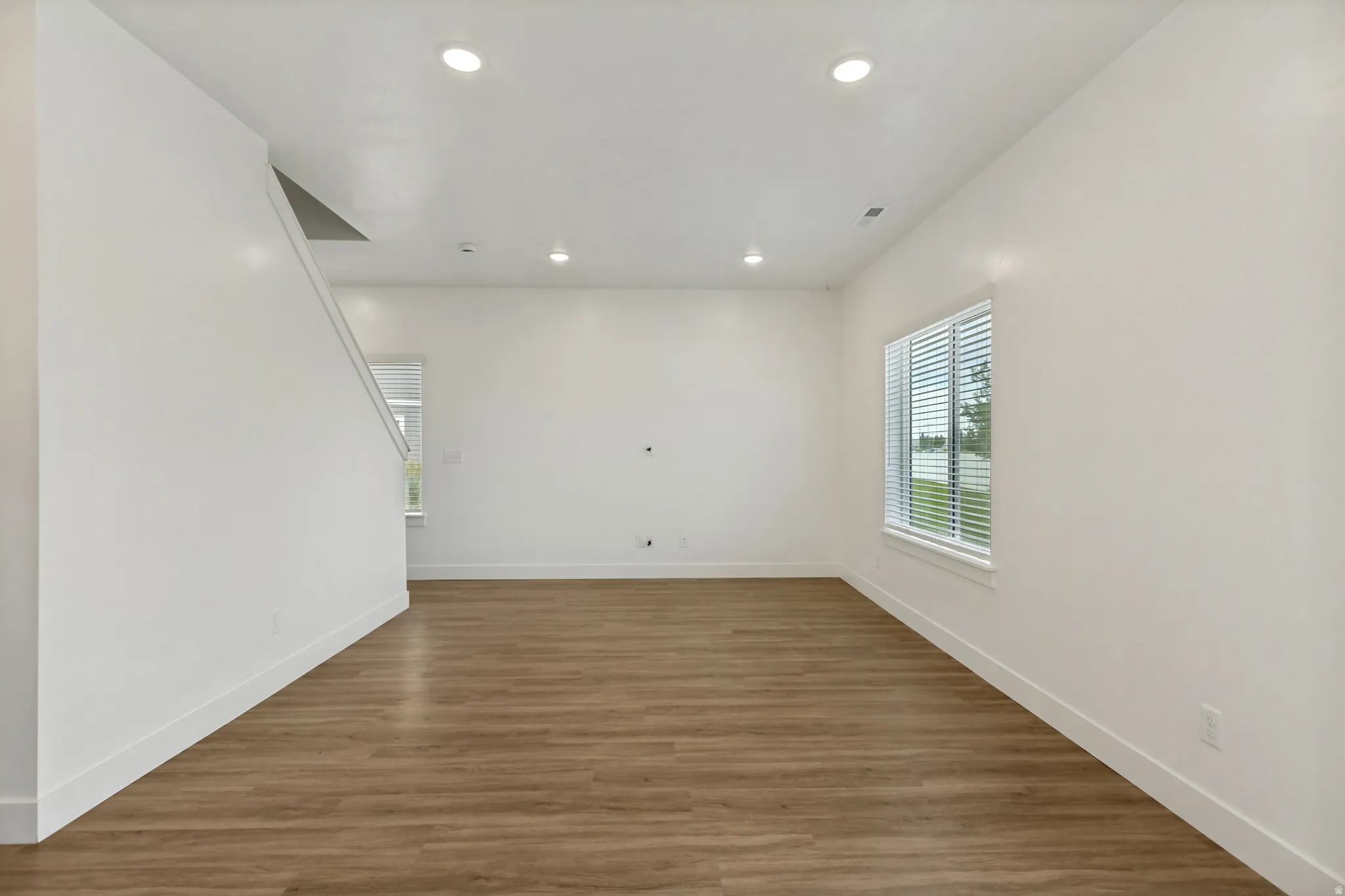 Spacious room with wood-finish flooring, white walls, recessed lighting, and a large window with blinds bringing in natural light