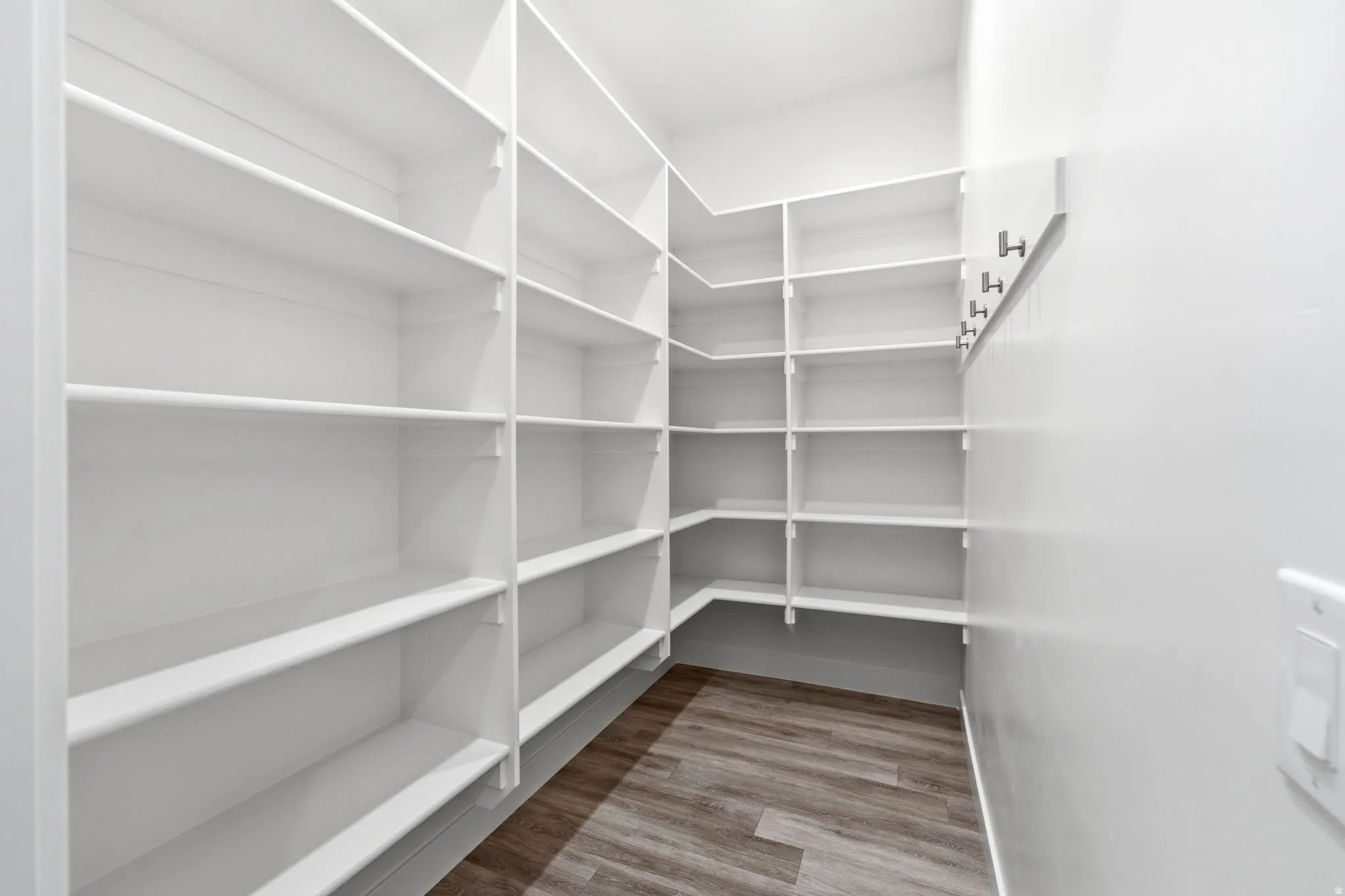 Spacious walk-in pantry featuring white shelving, light wood-finish flooring, and wall-mounted hooks