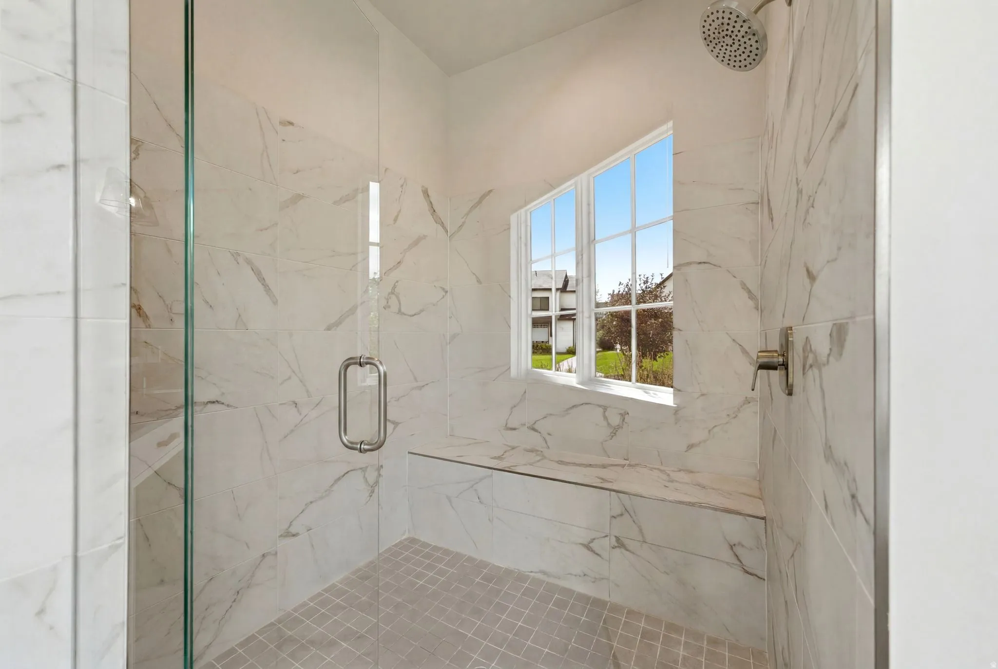 Walk-in shower featuring a glass enclosure with a metal handle, white marble-look wall tiling, a built-in bench, a rain shower head, and a grid-patterned window