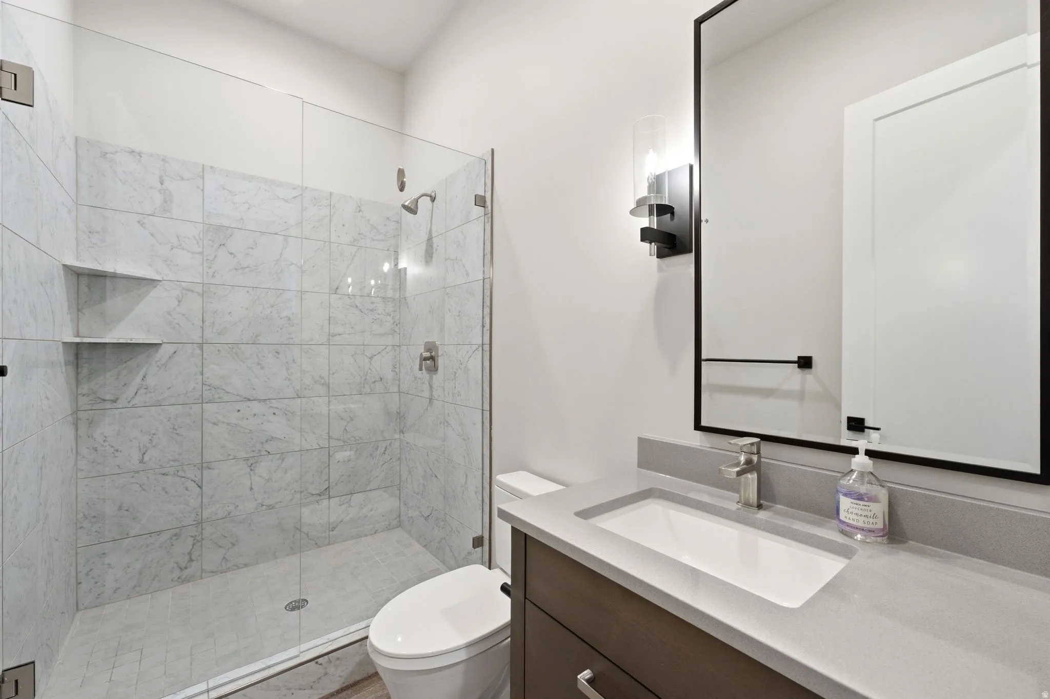 Bathroom featuring a glass-enclosed shower with tiled walls and built-in shelving, a contemporary vanity with an integrated sink and light grey countertop, and a sleek mirror with a matte black frame