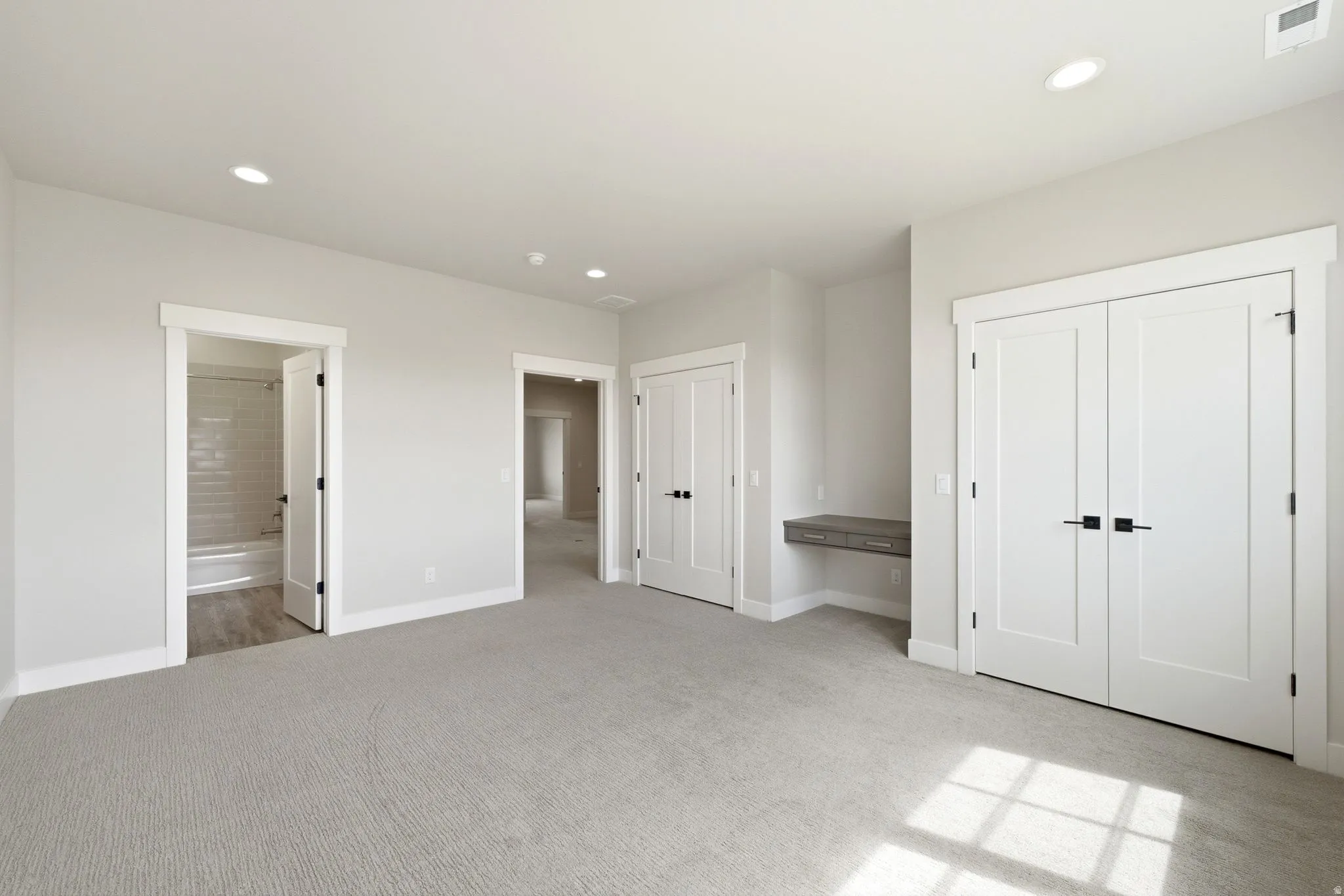 Spacious room featuring light gray carpeting, recessed lighting, white trim, a built-in floating desk, and multiple white panel doors with matte black hardware
