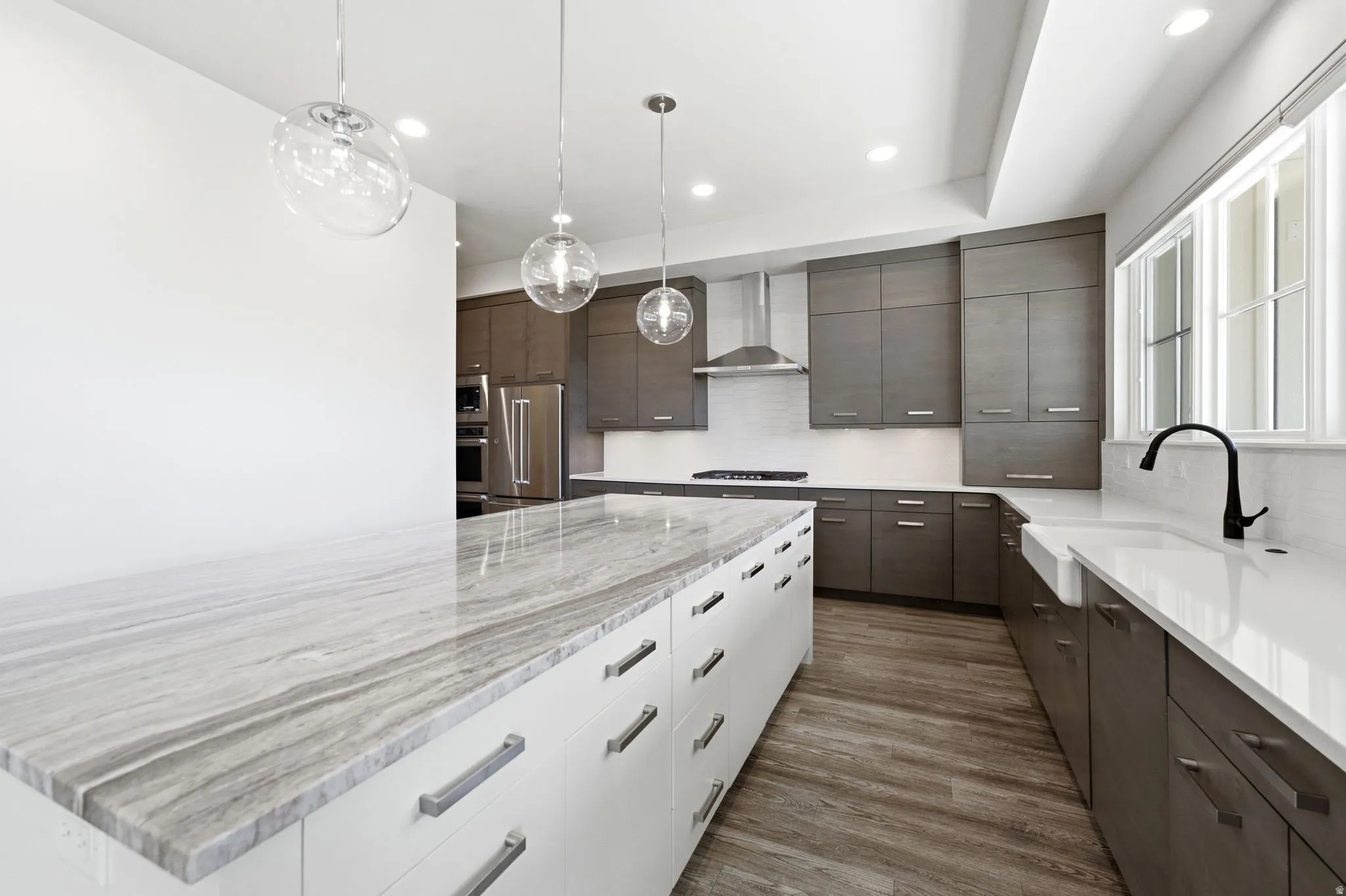 Contemporary kitchen featuring a stone-top island with extensive drawer storage, wood-finish flooring, dark-toned cabinetry, and a white farmhouse sink with a matte black faucet