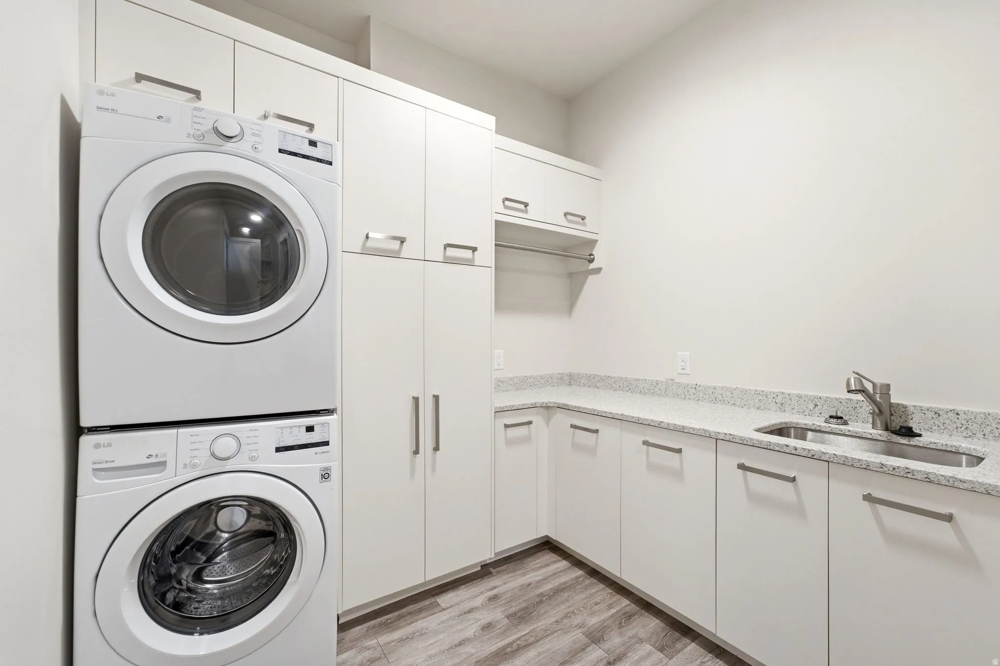 Dedicated laundry area featuring stacked front-loading appliances, extensive white cabinetry, a built-in sink, light speckled countertops, and wood-finish flooring