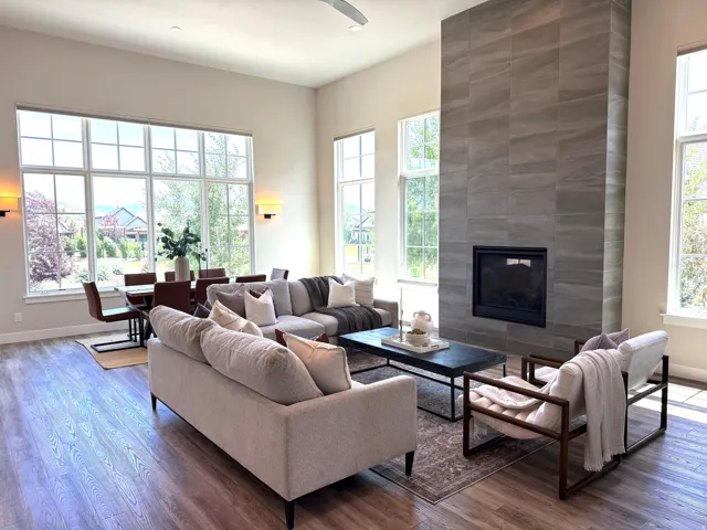 Expansive living area with high ceilings, large multi-pane windows, wood-finish flooring, a floor-to-ceiling tiled fireplace, and wall-mounted sconces