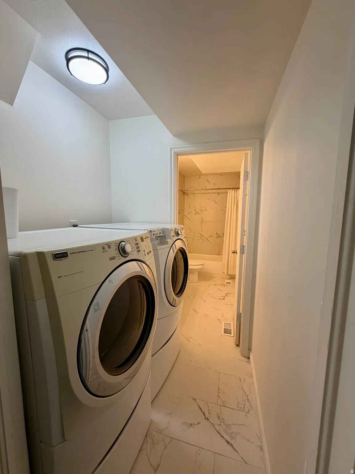 Dedicated laundry area featuring a front-loading washer and dryer, contemporary flush-mount ceiling light, and marble-look floor tiling extending into the adjacent bathroom